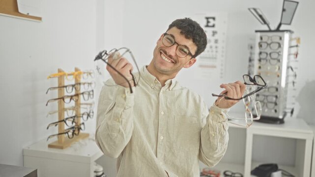 Man smiling while holding multiple eyeglass frames in both hands in an optician shop with eye chart and display racks visible; welcoming service.