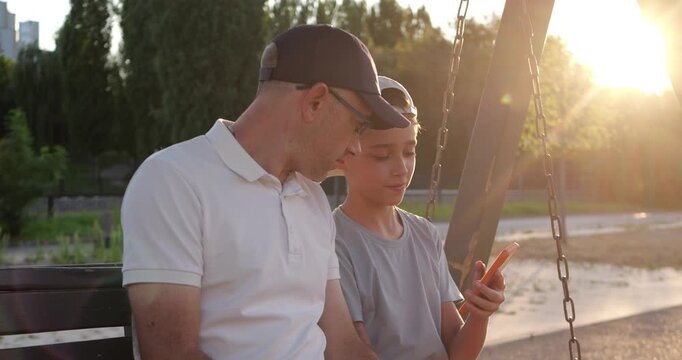 Dad and son use smartphone together on park swing at sunset. Family shares technology and conversation outdoors at sunset. Warm evening shows parenting, bonding and quality family time.