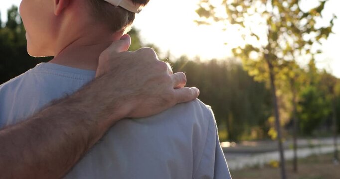 Protective father hand rests on young son's shoulder in park at sunset. Intimate family scene represents trust, guidance, reassurance and strong bond between generations together outdoors.