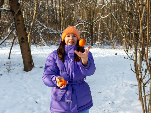Cheerful woman in purple winter jacket playfully tosses a fresh mandarin into the air while holding another citrus fruit in a snowy forest. Lifestyle concept for healthy snacking, vitamin-rich food, w