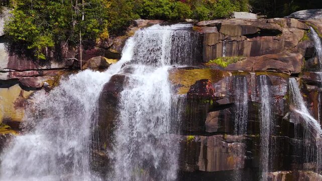 Peaceful mountain waterfall falls through rocky wilderness surrounded by yellow and red autumn leaves. Sunbeams illuminate mist rising from rushing water.