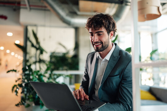 Young professional businessman smiling, actively using laptop for remote work in a modern office environment