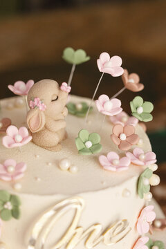 Close-up of first birthday cake decorated with small sleeping bunny figurine and delicate pastel sugar flowers on smooth white buttercream icing, pearl sugar accents and piped gold script visible at
