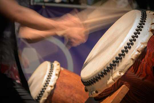 A group of taiko drummers performs outdoors, striking traditional Japanese drums with wooden bachi sticks in a coordinated, energetic ensemble performance.