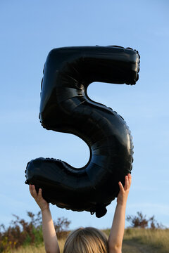 Child holding a black number five balloon against blue sky