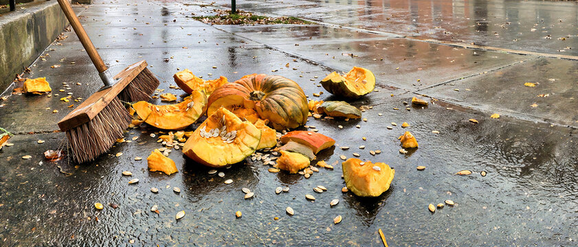 Smashed orange pumpkin with scattered seeds lying beside wooden push broom on wet pavement street. Post Halloween cleanup of broken gourd on rainy city sidewalk during autumn season