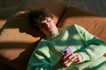 Obraz premium Young man with curly hair lying on sofa checking smartphone in mint sweater for relaxed lifestyle and social media projects