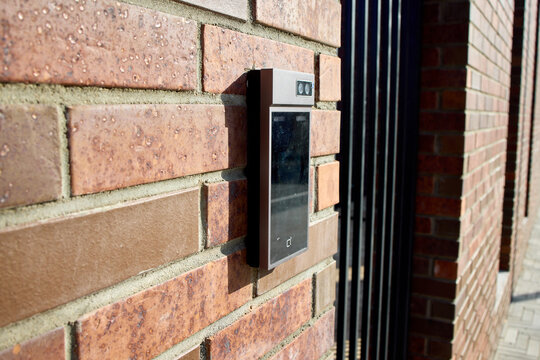 Smart doorbell camera mounted on red brick exterior wall beside residential front door in daytime, showing illuminated ring button, lens, and security monitoring