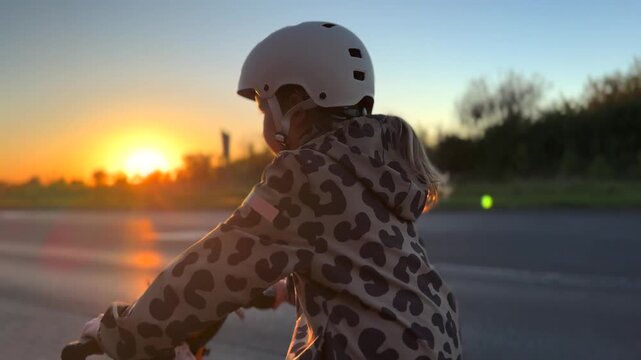 Child rides bicycle along quiet road at sunset, wearing white helmet and leopard print hoodie. Warm light glows on pavement while dark trees line horizon behind moving rider, with ponytail visible.