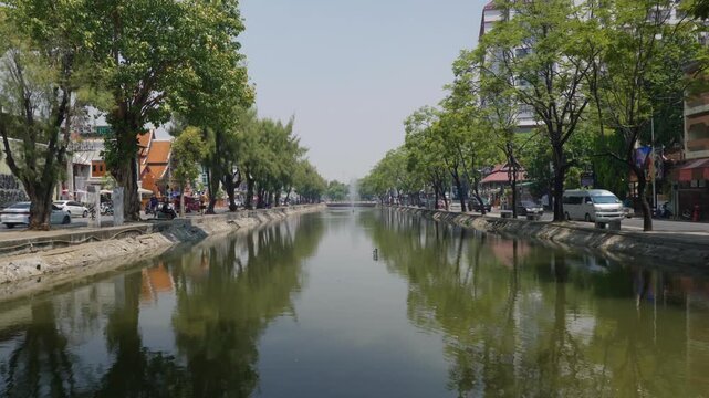 Wide shot along the historic city moat canal lined with lush green trees and street traffic under hazy, polluted skies during crop burning season in Chiang Mai, Northern Thailand.