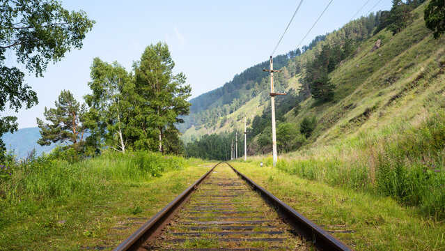 Empty railway track stretching into the distance through a green summer landscape, Lake Baikal, Russia. Historical rural train line with mountains and trees. Journey and travel concept