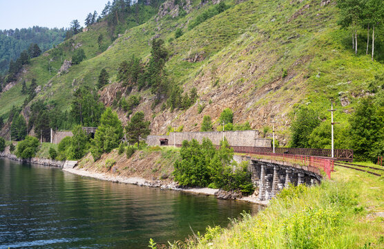 Single track railway at Lake Baikal, Russia. Old stone bridge and tunnel built into a steep green mountain. Summer travel, landscape and railroad infrastructure