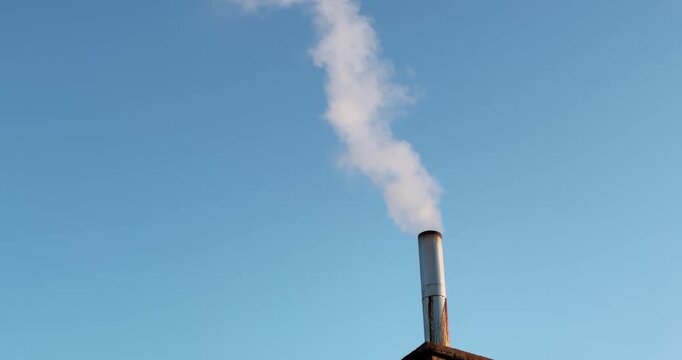 A chimney is smoking and the sky is blue. The chimney is tall and the smoke is coming out of it