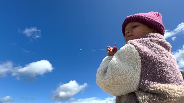 Child flying kite under bright blue sky while holding red string spool and looking upward. Warm fleece jacket and knit hat suggest cool weather as small kite drifts across open air above.