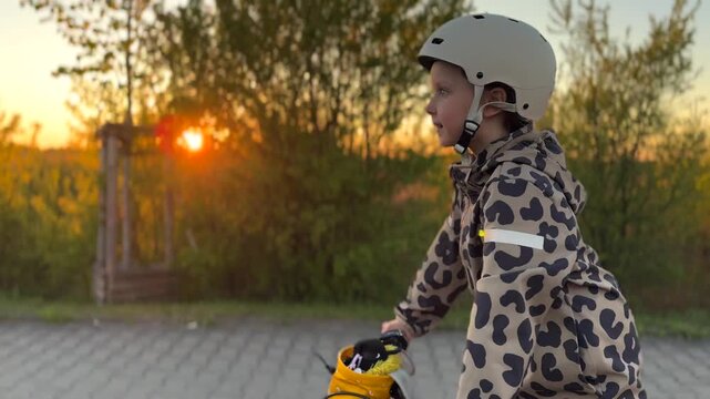 Child cyclist moves through evening light. White helmet, hooded patterned jacket, and upright riding posture stand out as young rider pedals along path near trees while sun sinks low behind horizon.