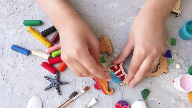 Top view of child's hands coloring small wooden striped lighthouse cutout with blue wax crayon, surrounded by various colorful crayons, art supplies, and sea-themed shapes on a textured gray surface