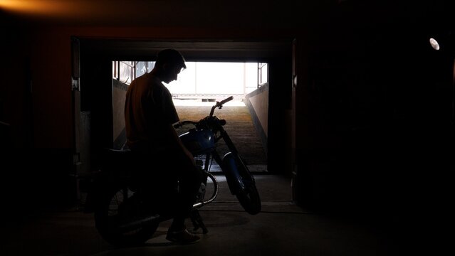 Man standing beside a vintage motorcycle in a dark garage, silhouetted against the bright light of the outdoor entrance, reflecting on travel and new beginnings