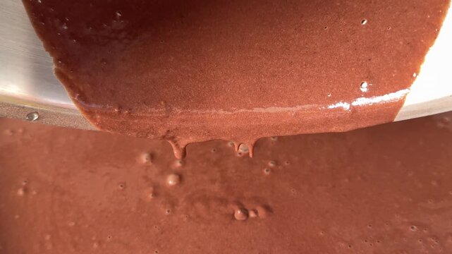 Thick chocolate mixture slowly falls in tiny drops into a baking pan, showing the rich glossy texture and consistency of freshly mixed cocoa batter. Close up.