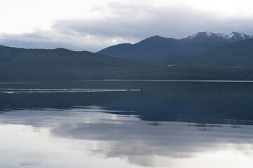 Serene mountain range mirrors on glassy lake as distant rowers glide beneath moody clouds, with mountain, lake, water, reflection detail, showing rower, rowboat, boat, calm context © Anna Ryzhkova