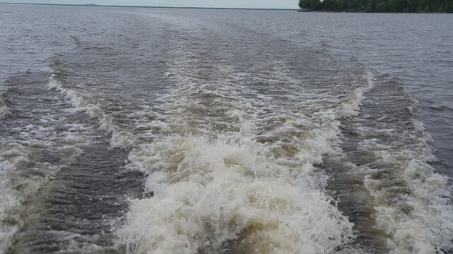 Rear view of a foamy wake spreading across a lake behind a moving motorboat, creating textured waves on the water surface.