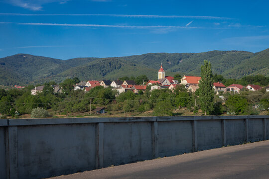 The village of Kostolna Ves in Slovakia nestles among red-roofed houses and a church spire, framed by forested mountains under a bright blue sky with contrails overhead.
