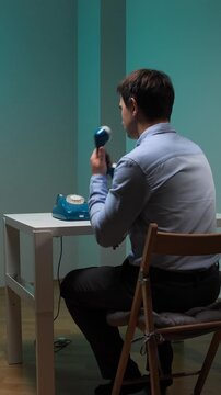 Vertical video young man sitting at a white desk, intently dialing a blue vintage rotary phone, symbolizing past communication methods, nostalgia, and a slower pace of life