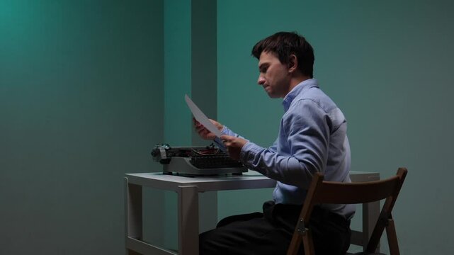 Man intensely reading a typed manuscript at a white table, holding paper beside a vintage typewriter on a teal background, focused on editing and creative work