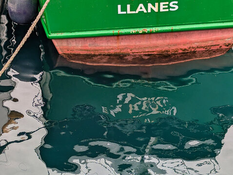 Close-up of the green stern of a fishing boat marked "LLANES," reflected in the still harbor water alongside a mooring rope and fender, Llanes, Asturias, Spain