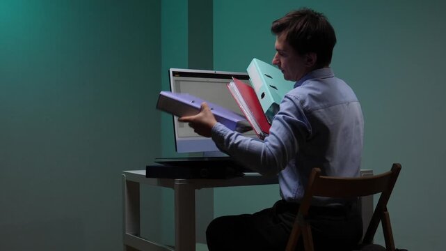 Busy man struggling with a stack of folders while working at a computer, representing the concept of administrative overload, stress, and heavy paperwork