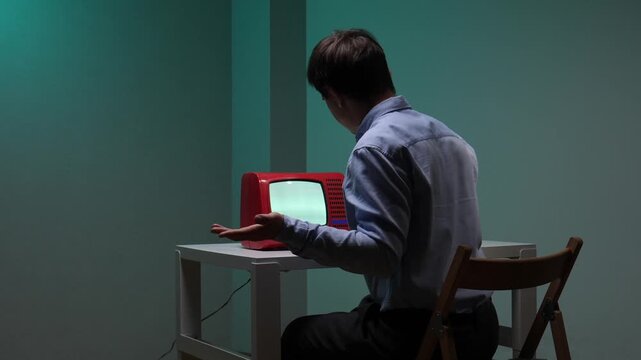 Young man sitting in a dim room watching a glowing red retro television, holding his hand near the blank screen, exploring themes of technology, nostalgia, and communication