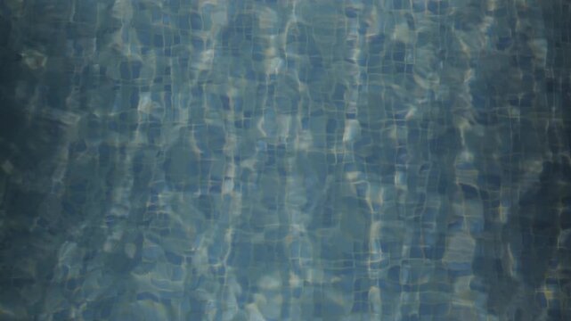 Close up of rippling swimming pool water with shimmering sunlight patterns across blue mosaic tiles creating an abstract aquatic background
