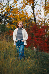 Obraz premium A young red-haired girl in a white jacket walks through a grassy field with colorful autumn foliage behind her.