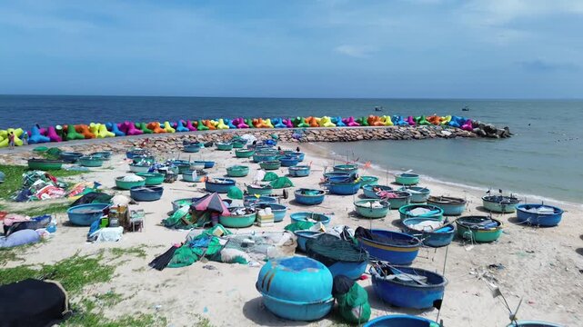 Low angle aerial view of traditional Vietnamese basket boats resting on sand and grass in Mui Ne. Colorful X-shaped wave breakers along the shoreline with sea in Background in Binh Thuan Vietnam.