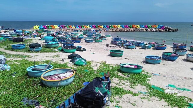 Low angle aerial view of traditional Vietnamese basket boats resting on sand and grass in Mui Ne. Colorful X-shaped wave breakers along the shoreline with sea in Background in Binh Thuan Vietnam.