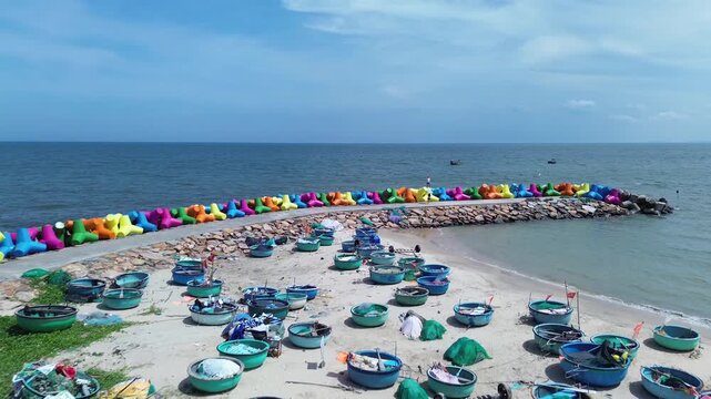 Low angle aerial view of traditional Vietnamese basket boats resting on sand and grass in Mui Ne. Colorful X-shaped wave breakers along the shoreline with sea in Background in Binh Thuan Vietnam.