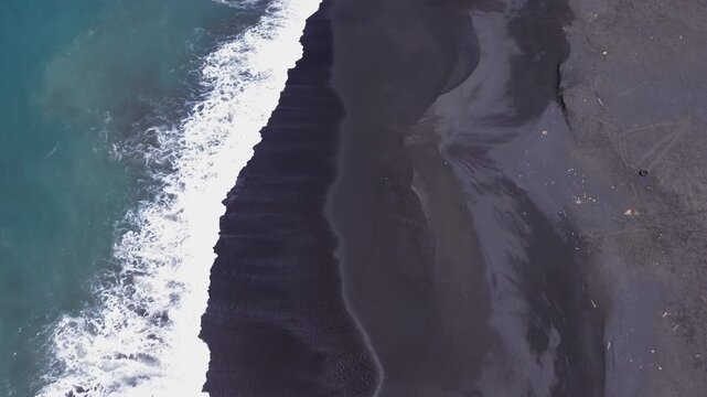 Birds eye top down aerial drone shot of white ocean wave foam crashing and flowing over black volcanic sand shoreline along Jumpai beach in Klungkung regency, Bali island, Indonesia.