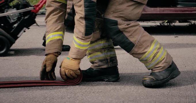 Firefighter team - fireman equipped with emergency services response firefighting crew uniform and gear reeling in fire hose. Department crew from fire station and car wreckage is in scene background.