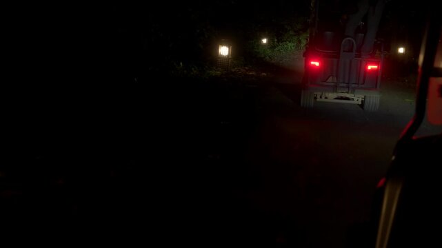 Low viewpoint from a moving electric buggy follows another vehicle along a dark rainforest road, with red tail lights and path lights visible ahead.