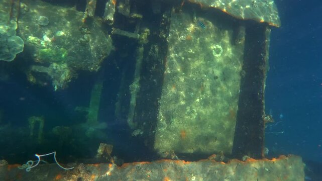 Camera looks down into an underwater shipwreck opening, revealing rusted structure, coral growth and shifting sunlight reflections in clear blue water.
