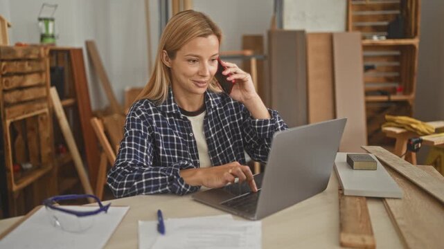 Woman carpenter talking on smartphone and typing on laptop in workshop building, surrounded by planks and a workbench; practical focus.