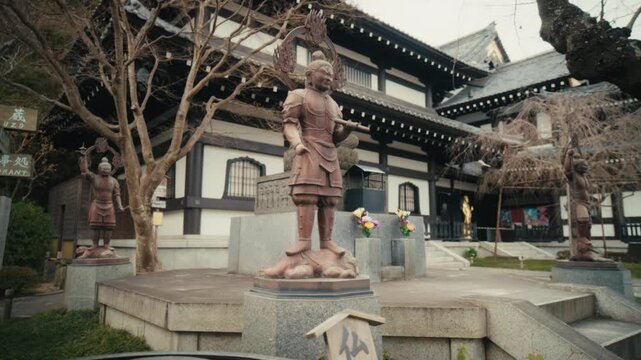 Weathered bronze guardian statues stand watch in the courtyard of Hasedera Temple in Kamakura, Japan, framed by the temple's traditional wooden main hall and surrounding bare trees.