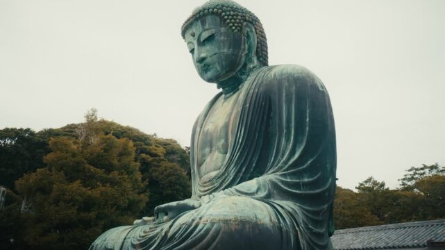A slow tilt shot captures the imposing bronze Great Buddha of Kamakura at Kotoku-in Temple, Japan, revealing its weathered patina and monumental scale against a wooded hillside backdrop.