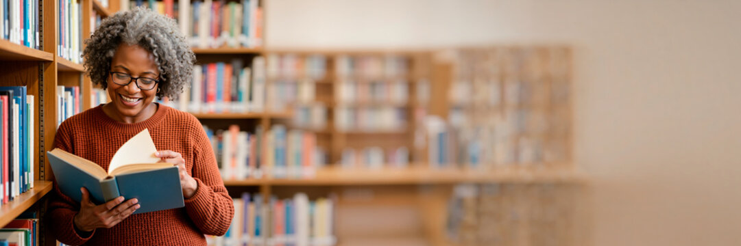 Smiling senior Black woman reading a book in a library. Happy mature African American female with glasses choosing a novel. Wide banner format with copy space for text