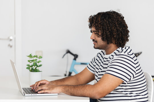 Young adult Indian man typing on laptop in side view at home office. Focused digital work session in minimalist workspace. Technology concept.