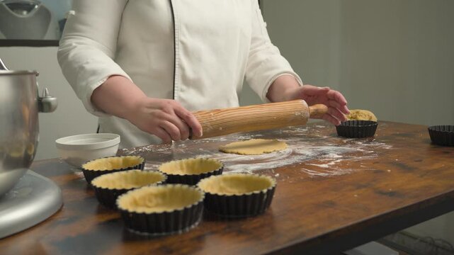 Pastry chef rolling out dough for tartlets on a floured wooden work surface, with several tart molds prepared for baking. Close-up of traditional pastry preparation in a bakery kitchen.