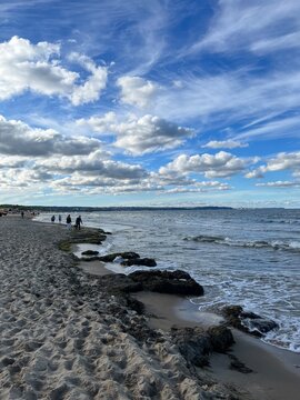 Scenic view of the Baltic Sea coast in Gdansk, Poland under a blue sky with white clouds. Sandy beach with seaweeds, rocks, and rolling waves on a windy fall day.