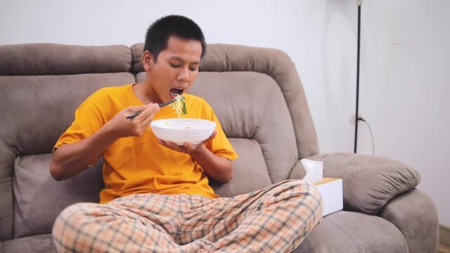 Young man eating noodles from bowl with chopsticks while sitting crossed on comfortable sofa