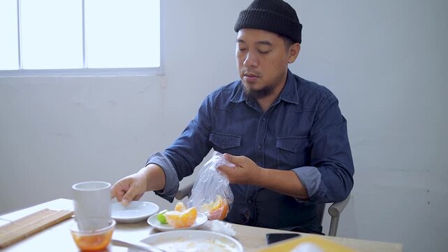 Asian man packs fruit peels into plastic bag and takes sip from his mug at table