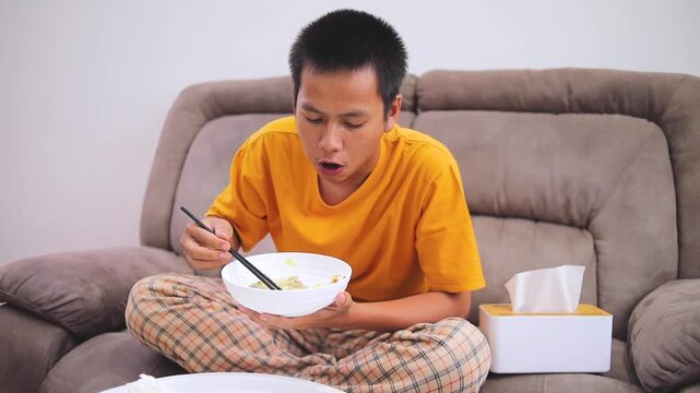 Young man eating chili on noodles with chopsticks and drinking from mug on cozy sofa at home