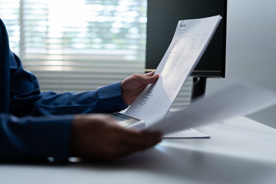 Businessman reviewing an invoice and financial documents at an office computer, representing accounting, billing, payment management, expenses, bookkeeping, and corporate finance workflow.